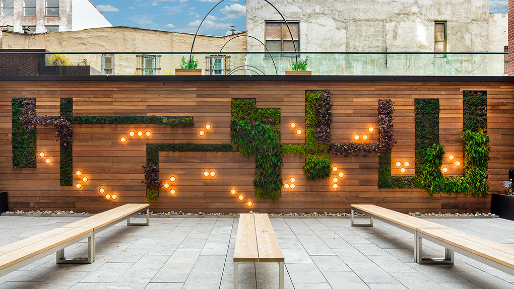 Fifth & Wythe Outdoor Courtyard with Tiled Flooring, Wood Panel Wall with Inset Greenery and Wood Bench Seating
